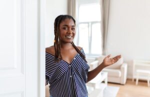 Happy young woman standing in doorway of modern apartment showing living room with hand.